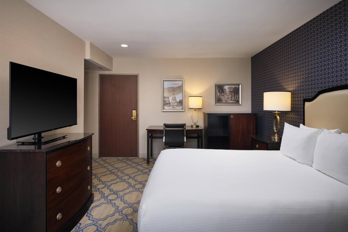 Une chambre d’hôtel avec un grand lit, un téléviseur posé sur une commode, un bureau avec une chaise et deux lampes fixées au mur.