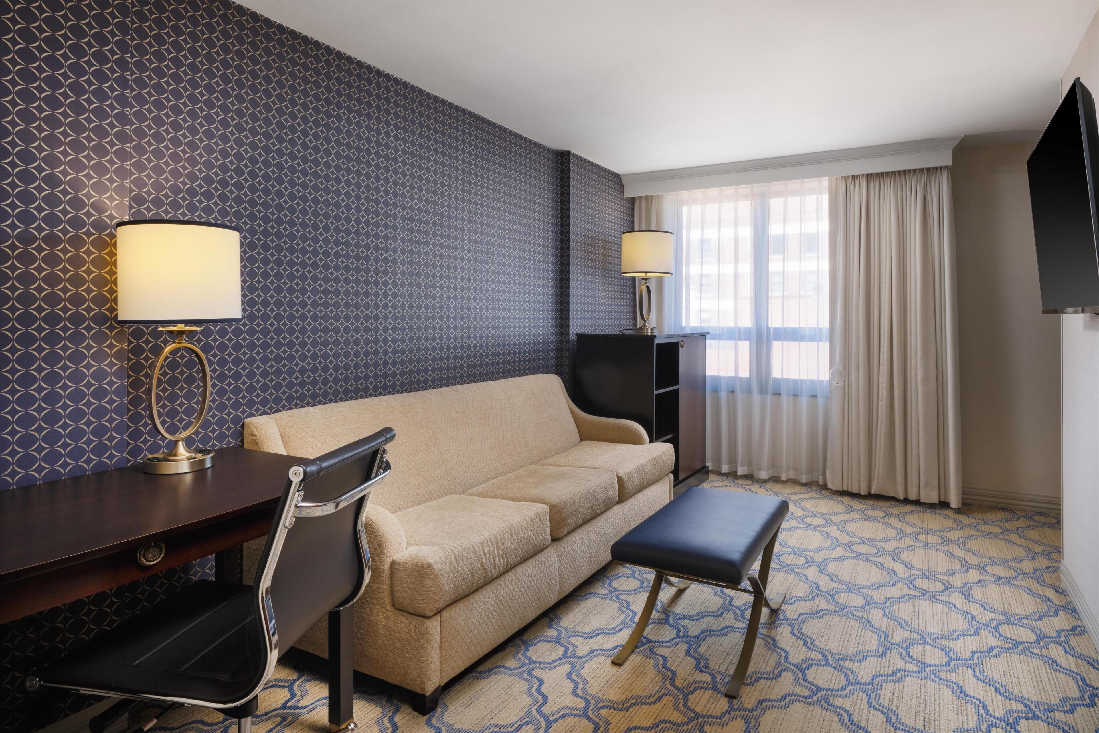 A hotel room with a patterned wall, a sofa, a desk with a lamp, a chair, a small table, and a TV.