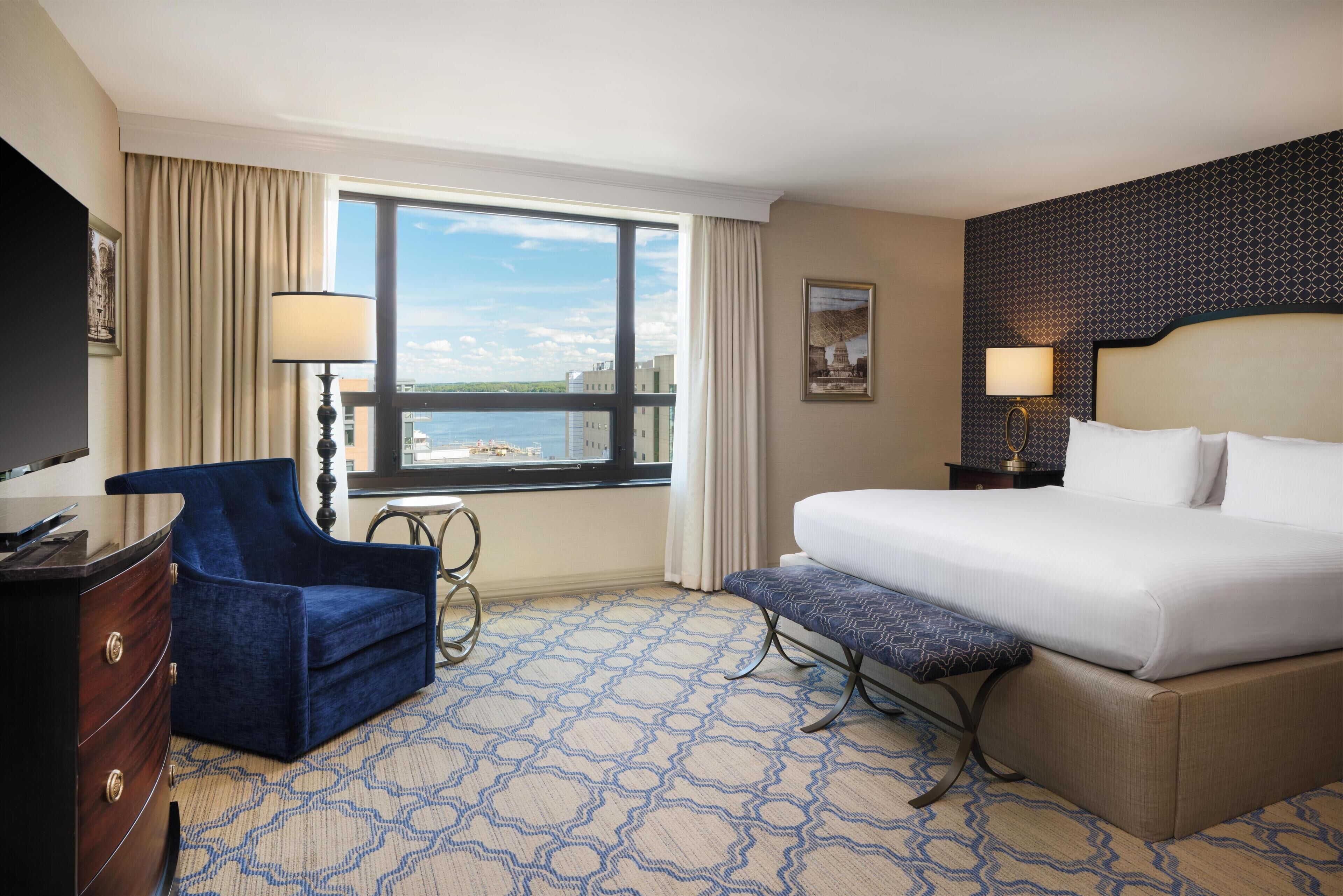 A hotel room with a large bed, a chair, a desk, a lamp, a window with a view, and a patterned carpet.