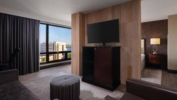 Quarto de hotel moderno com uma grande janela, televisão de tela plana fixada numa parede de painel de madeira, um sofá, um banquinho listrado e uma pequena mesa de cabeceira com uma luminária.