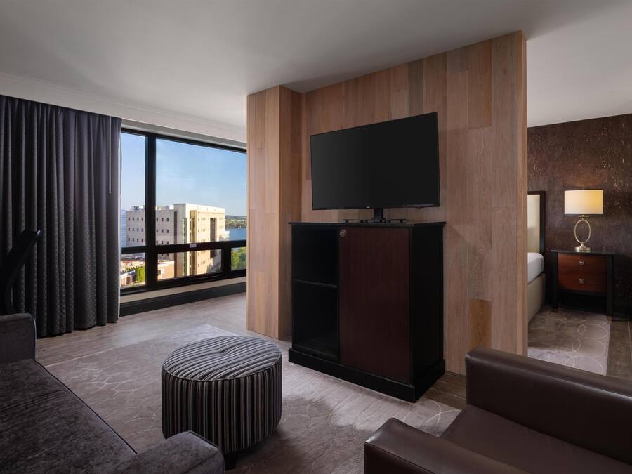 Quarto de hotel moderno com uma grande janela, televisão de tela plana fixada numa parede de painel de madeira, um sofá, um banquinho listrado e uma pequena mesa de cabeceira com uma luminária.