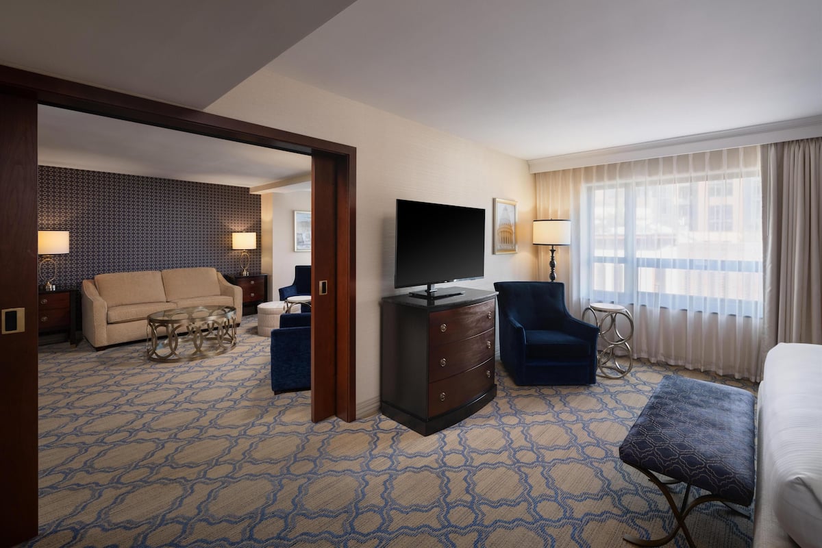 Une chambre d’hôtel équipée d’un canapé, d’une télévision, d’un bureau, d’une chaise, d’une petite table et d’une fenêtre avec des rideaux.