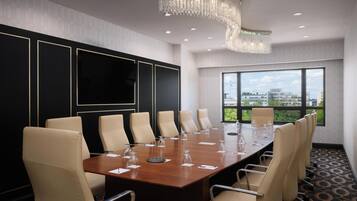 A conference room with a large table, beige chairs, a TV screen, and a view of buildings through large windows.
