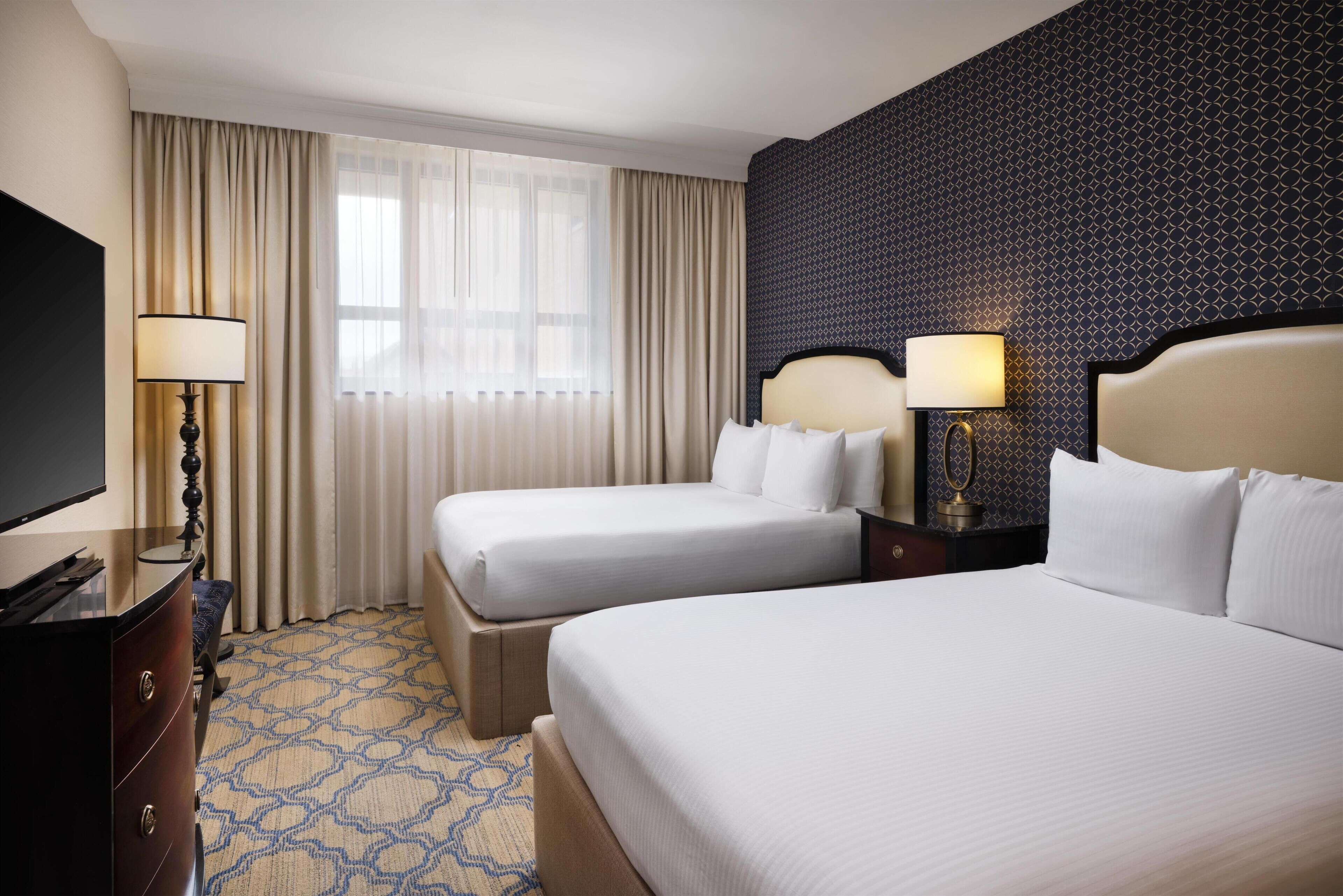 A hotel room with two beds, a TV, a desk, and a patterned wall.