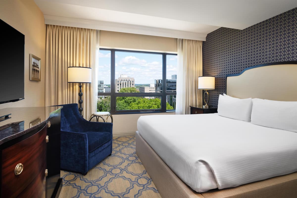 Une chambre d’hôtel avec un grand lit, un bureau avec une télévision, une chaise et une vue sur la ville grâce à de grandes fenêtres.