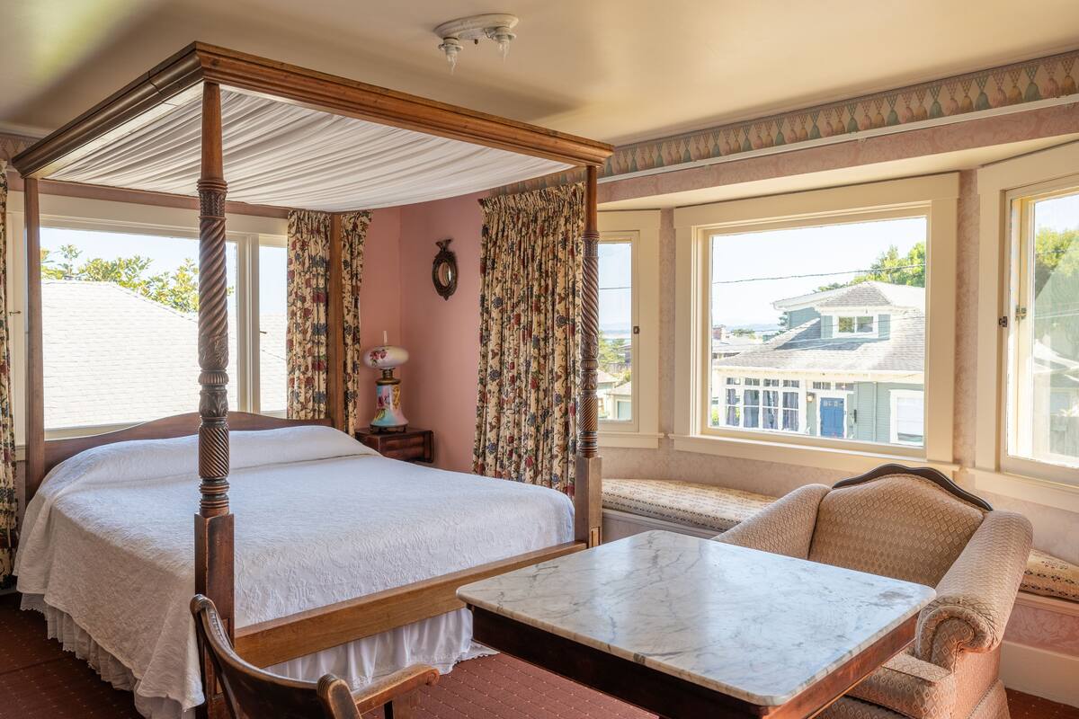 Ein Bett mit Baldachin, ein Marmortisch und eine gepolsterte Bank in einem Zimmer mit großen Fenstern und Blick auf Häuser.