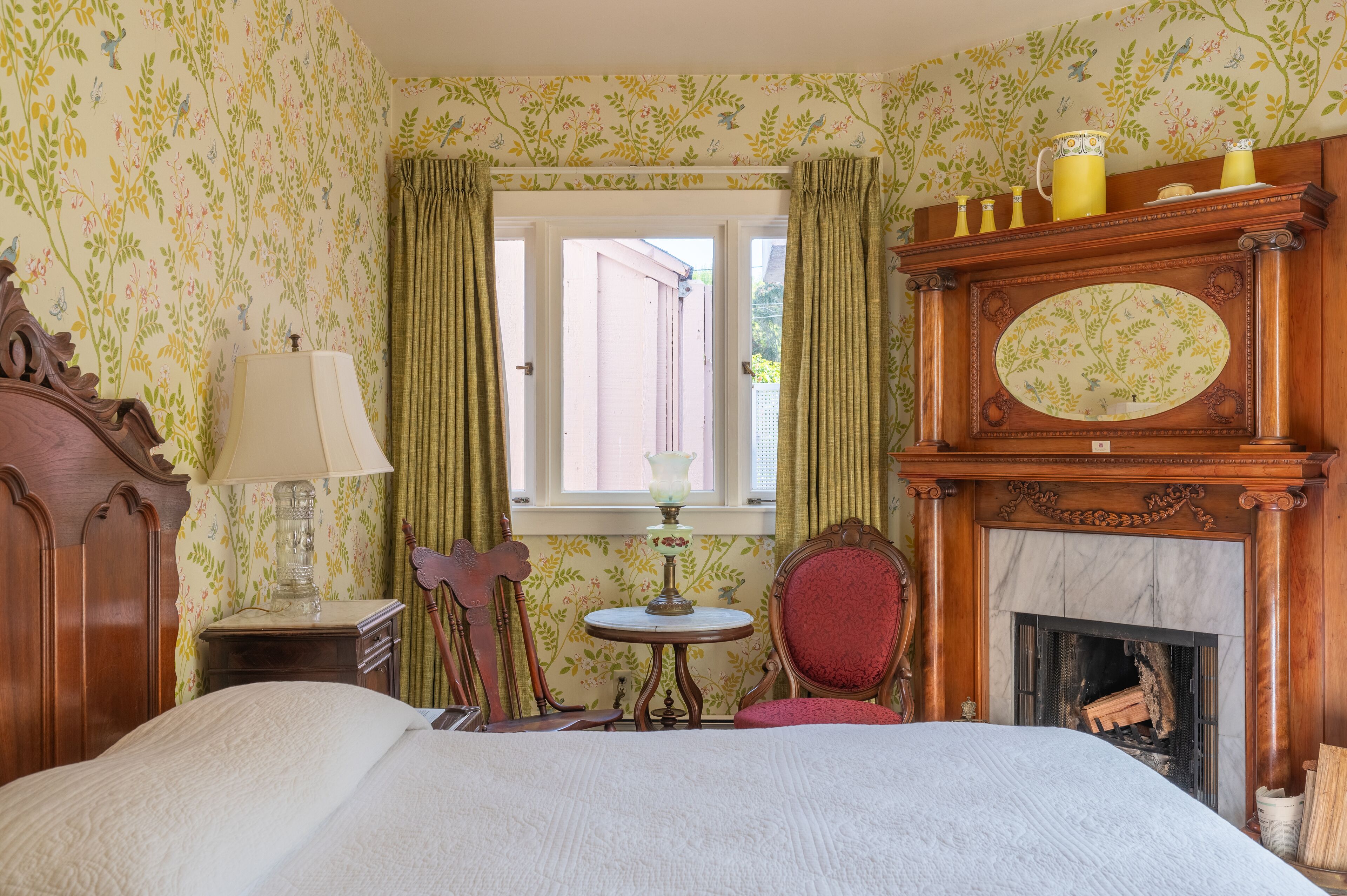 A bedroom with a bed, a wooden dresser, a table with a lamp, a chair, a fireplace, and floral wallpaper.
