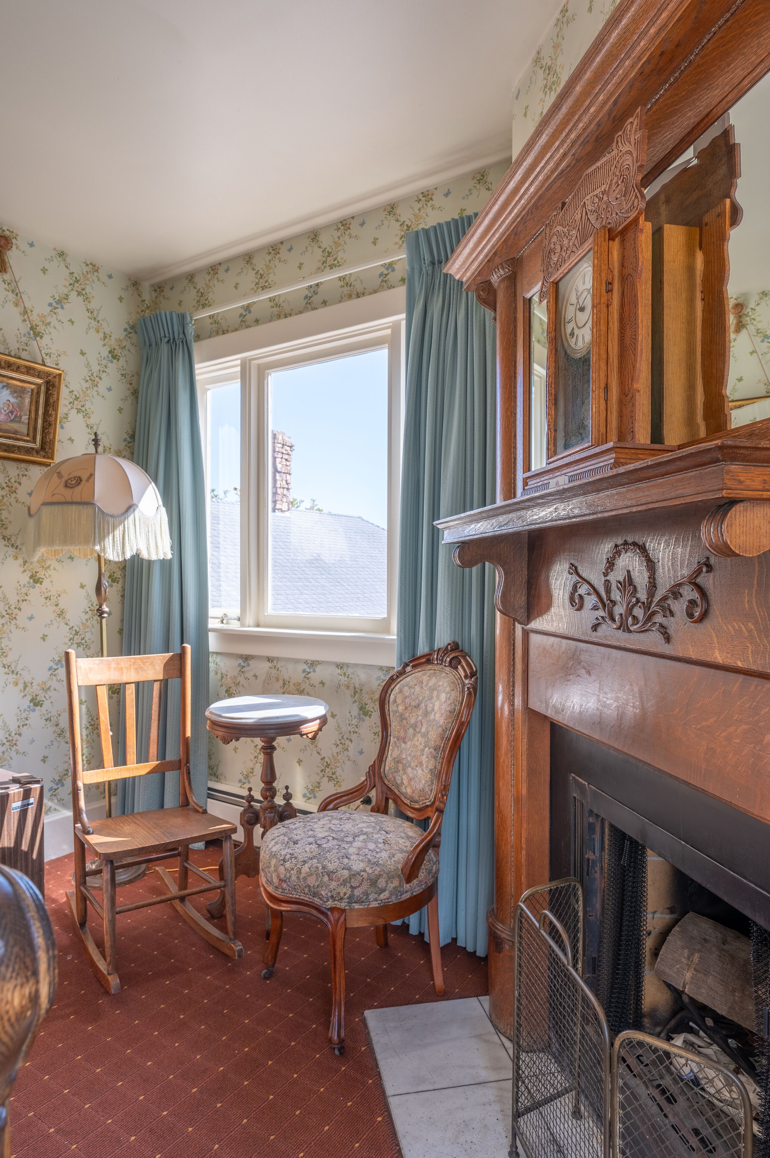 Quarto com lareira, duas cadeiras de madeira, uma mesinha e janela com cortinas.