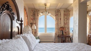 A bedroom with a large bed, ornate headboard, and a view of the sea through a window with floral curtains. - Martine Inn (Pacific Grove)
