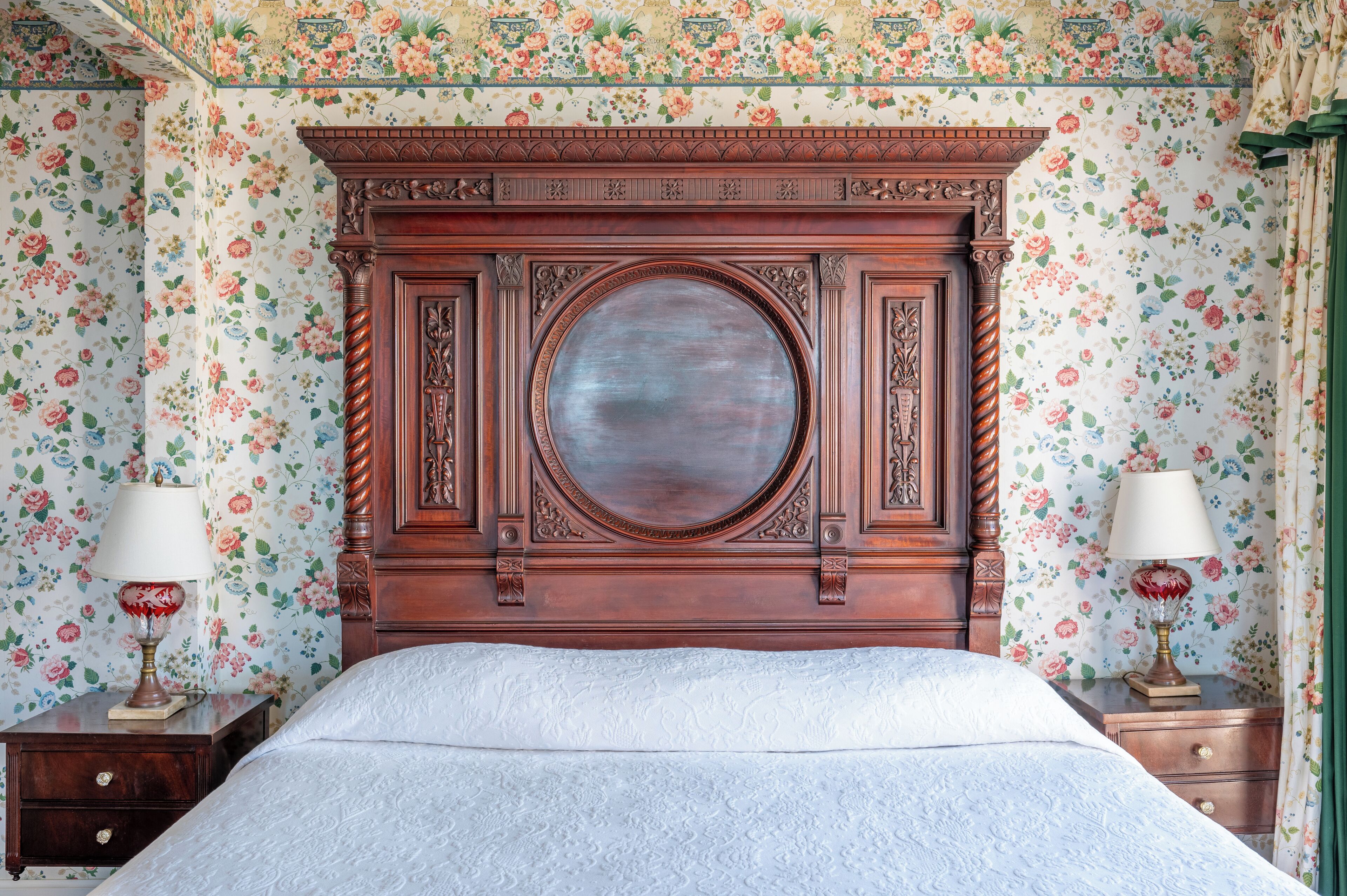 A bedroom with a wooden headboard, floral wallpaper, a round mirror, and two bedside tables with lamps.