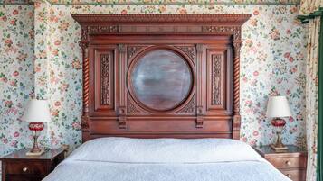 A bedroom with a wooden headboard, floral wallpaper, a round mirror, and two bedside tables with lamps.