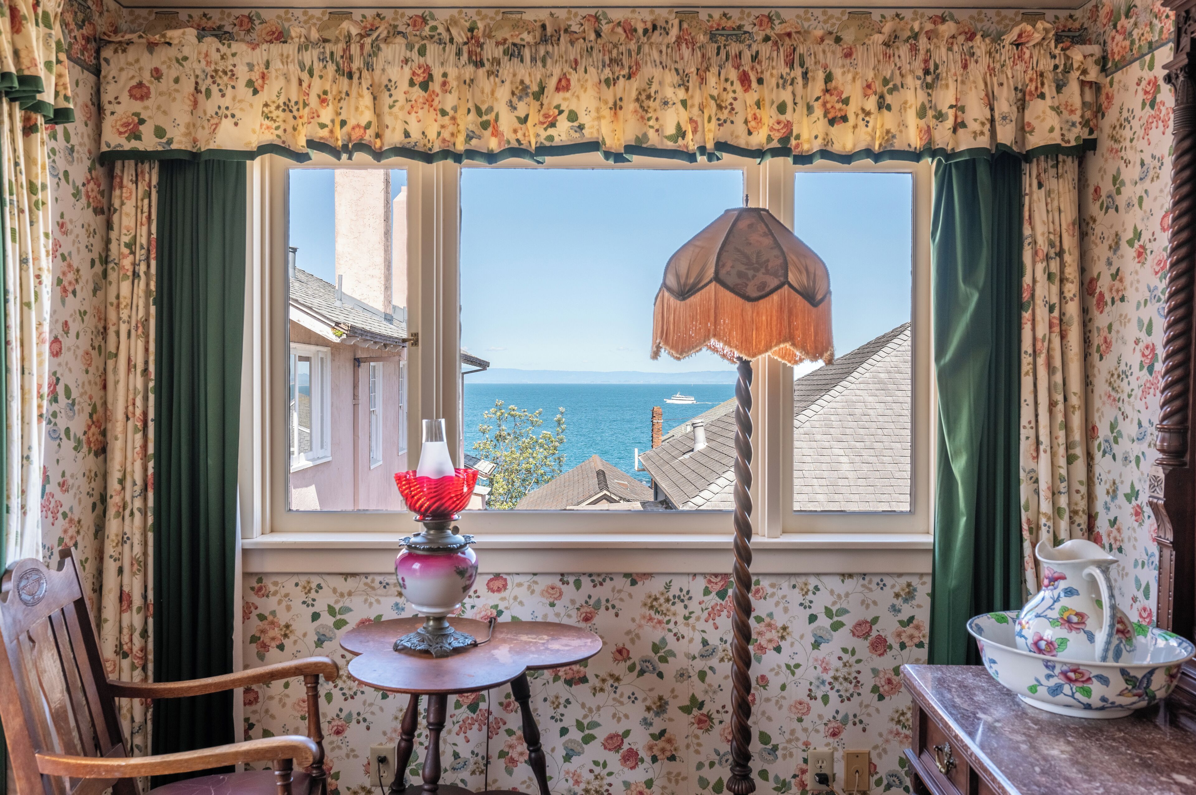A cozy room with a view of the sea, a table with a lamp, and floral wallpaper.