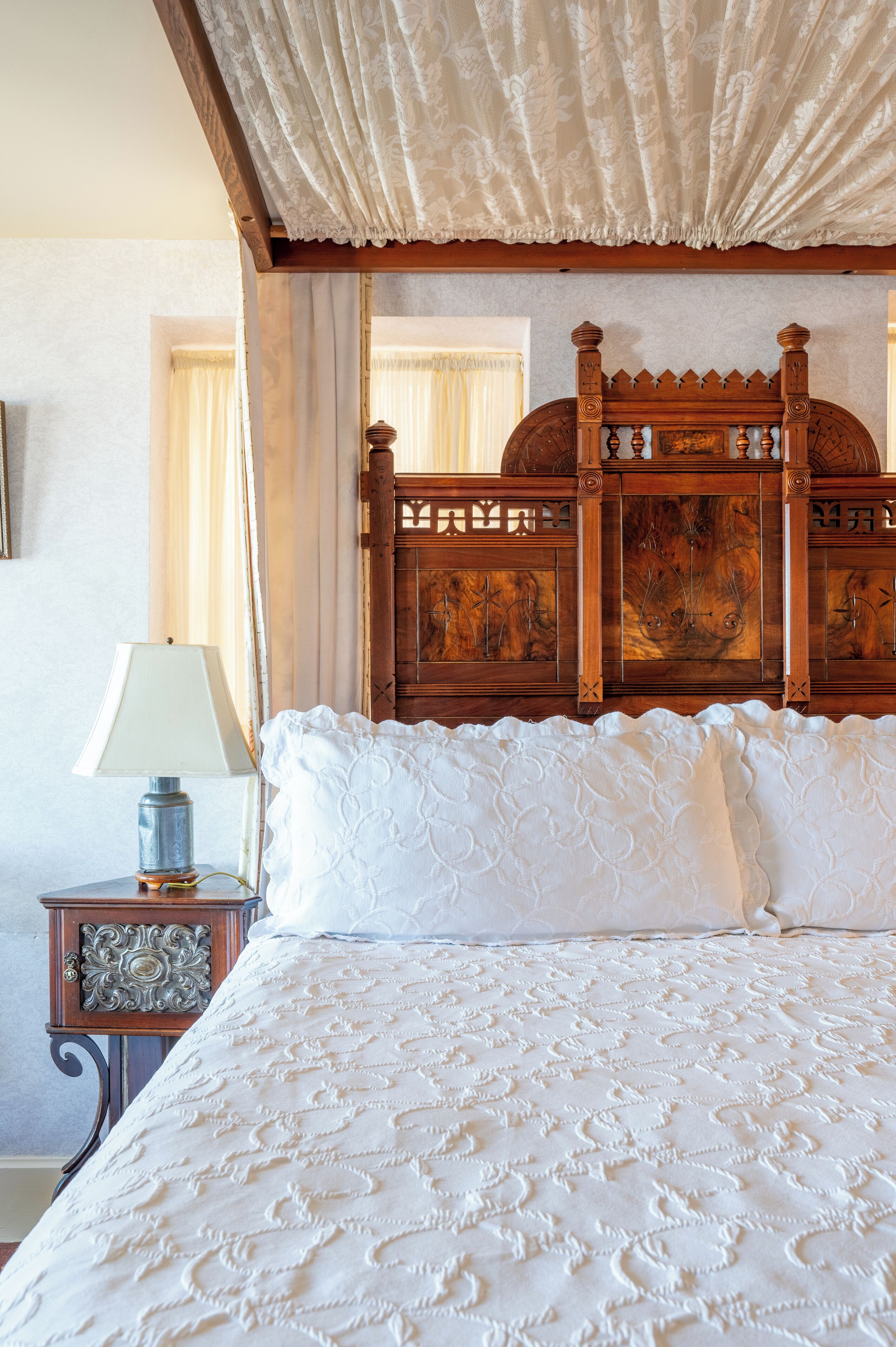 A bed with a white quilt, a wooden headboard with carvings, and a nightstand with a lamp.