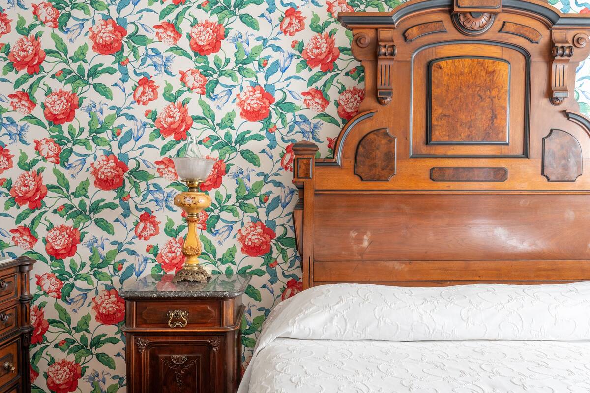 Ein Schlafzimmer mit blumigem Wandpapier, einem hölzernen Kopfteil, einem Nachttisch mit Lampe und einem Bett mit weißer Bettdecke.