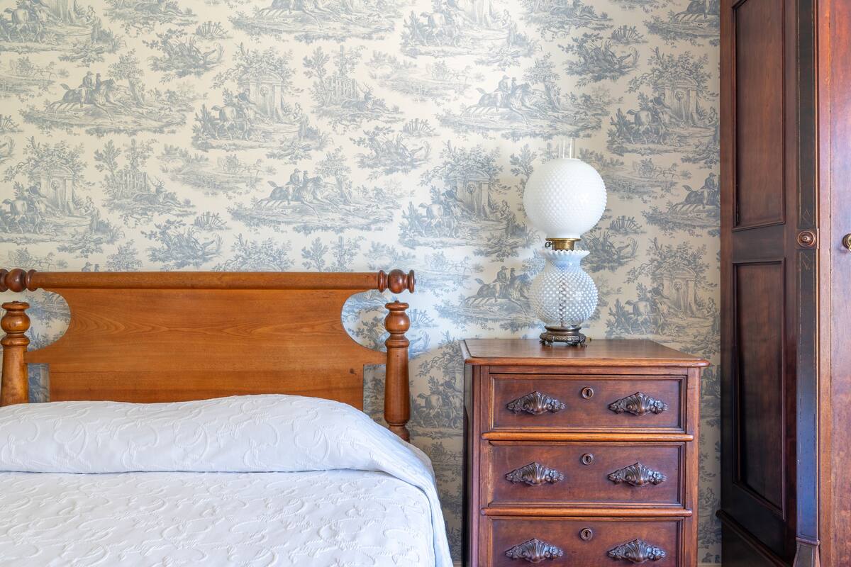 Ein Schlafzimmer mit einem hölzernen Kopfteil, einer weißen Bettdecke, einem Nachttisch mit Schubladen und einer Wand mit blumigem, blauem Tapetenmuster.