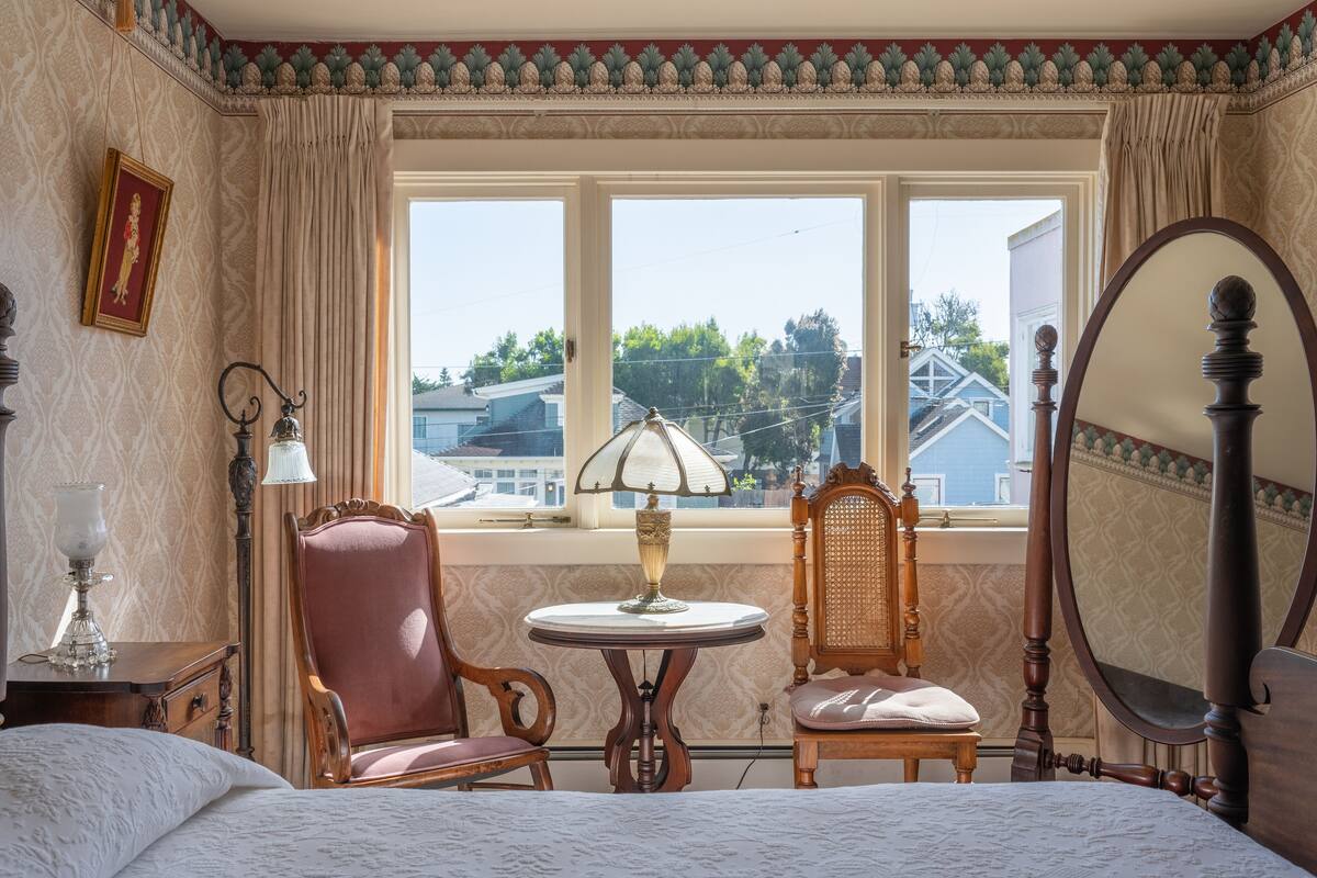 Ein Schlafzimmer mit einem Bett, zwei Stühlen, einem Tisch mit Lampe, einem Spiegel und einem Fenster mit Blick auf Häuser.
