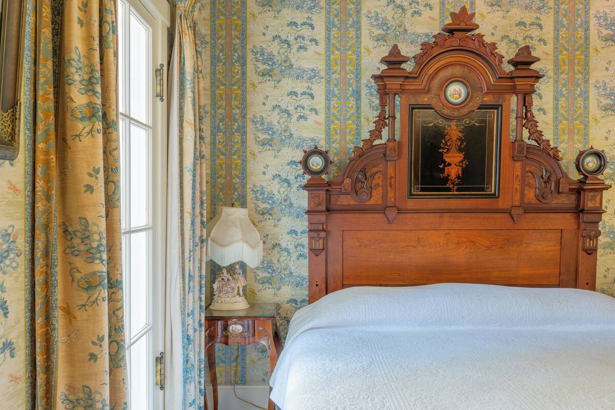 Ein Schlafzimmer mit einem hölzernen Kopfende, blumigem Tapetenmuster und einem Bett mit weißer Bettdecke.