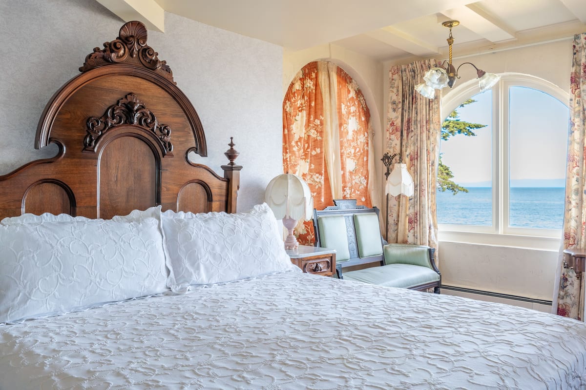 Ein Schlafzimmer mit einem großen Bett, einem Holz-Kopfteil, Meerblick durch ein Fenster und einem Sessel am Fenster.