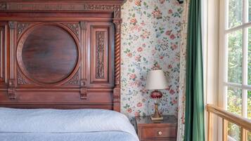Un dormitorio con un gran cabecero de madera, papel tapiz floral, una mesita de noche con lámpara y una ventana con vistas a la vegetación.