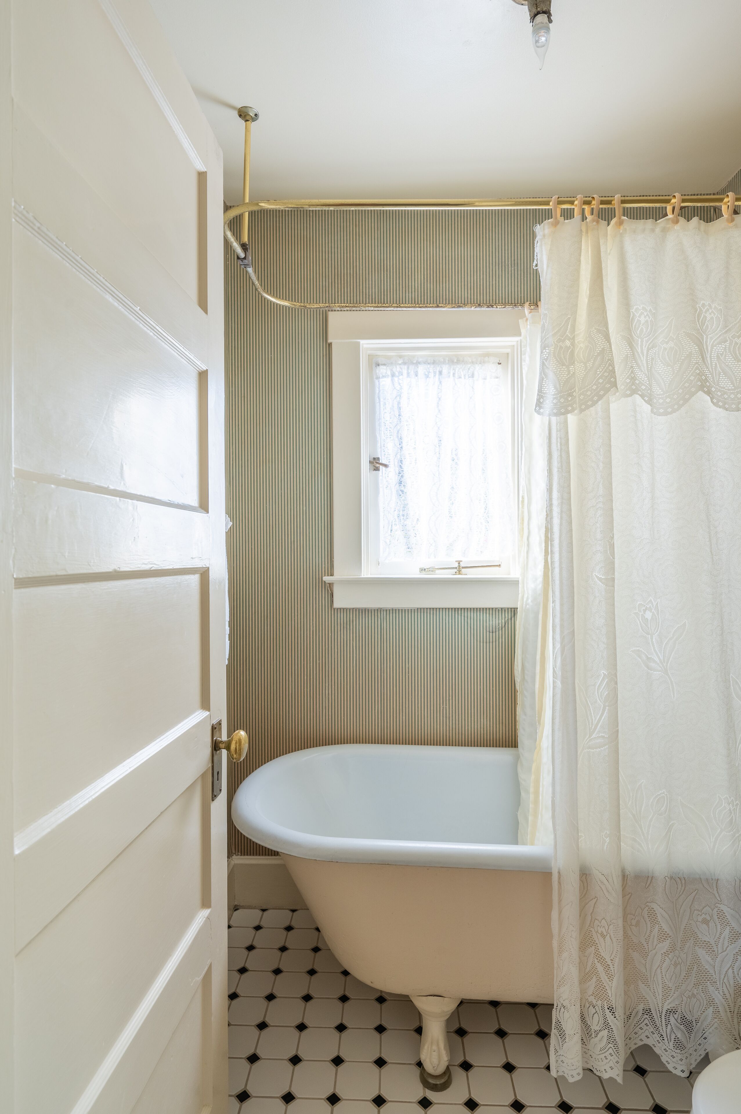A bathroom with a white bathtub, a window with a white curtain, and a tiled floor.