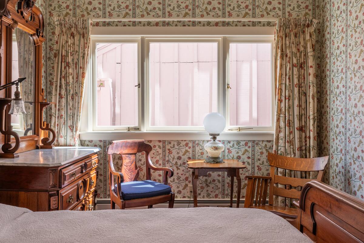 Ein Zimmer mit einem Bett, einem Holzschrank, einem Sessel, einem kleinen Tisch mit Lampe und einem Fenster mit Vorhängen.