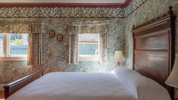 A bedroom with floral wallpaper, a wooden headboard, a bed with white linens, and a window with curtains.