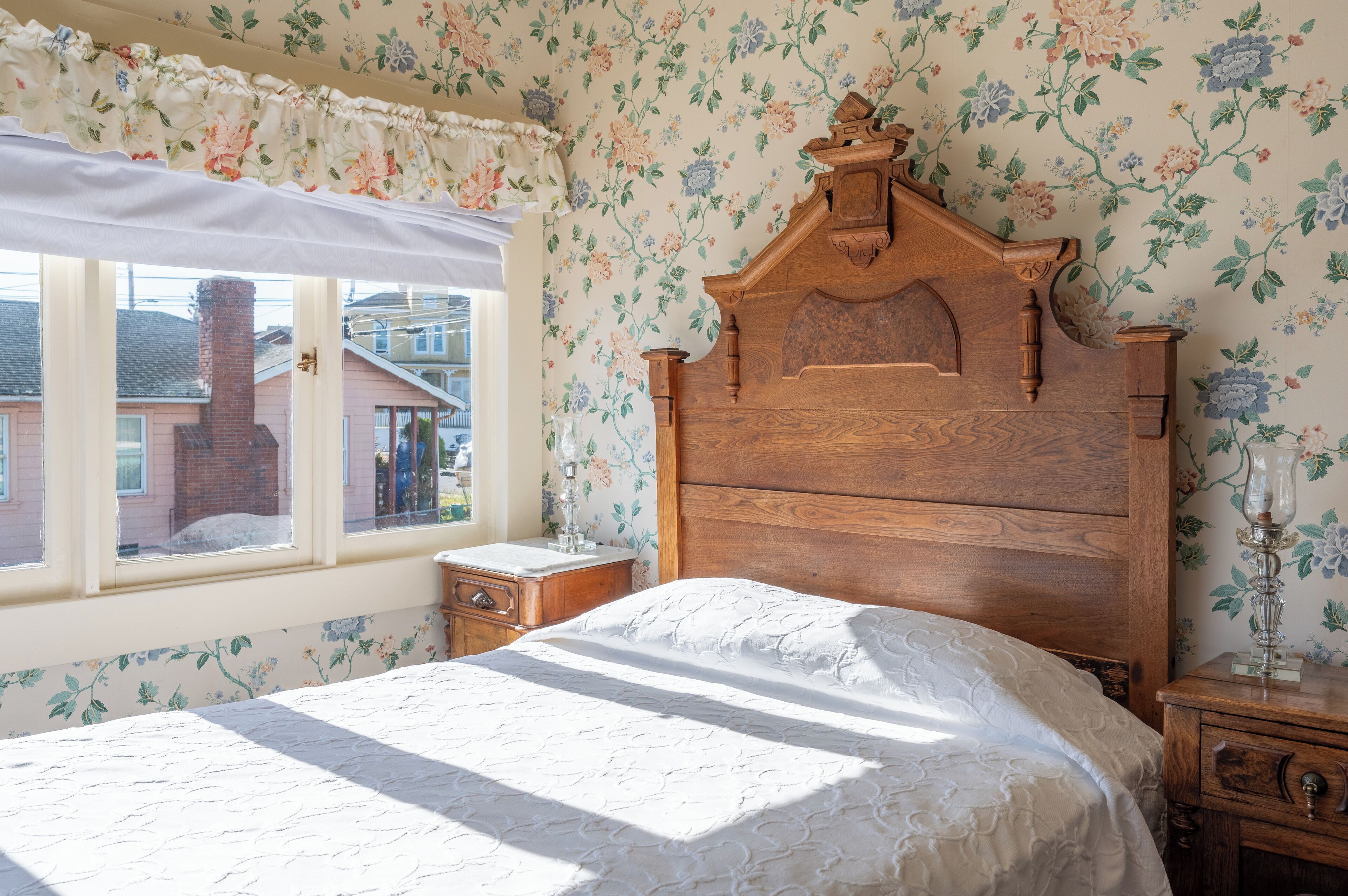 A bedroom with a wooden headboard, a floral wallpaper, a window with a view of a house, and a bedside table with a lamp.