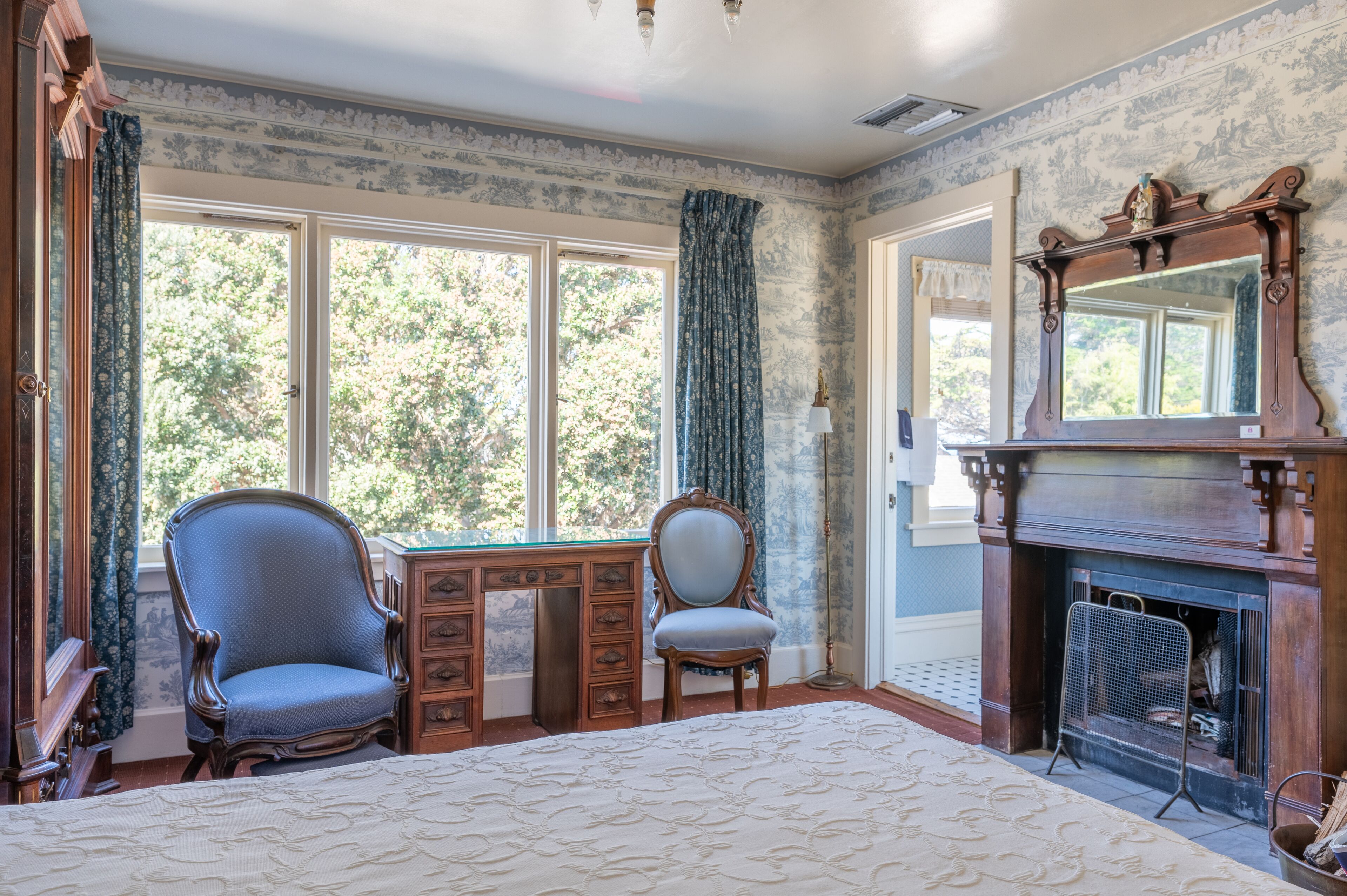 Une chambre dotée d’un grand lit, d’un bureau en bois, d’une chaise, d’une cheminée et d’une grande fenêtre avec des rideaux.