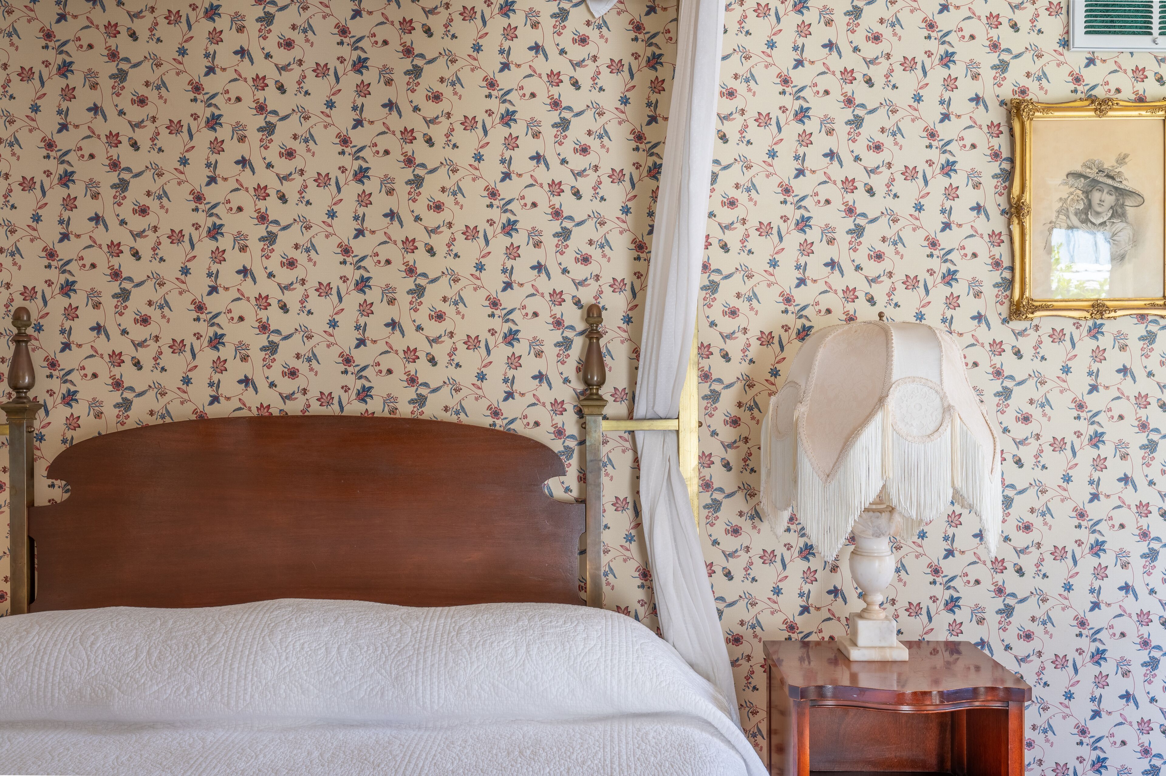 Une chambre à coucher avec un papier peint à motifs floraux, un lit en bois, une table de chevet avec une lampe et un tableau encadré au mur.