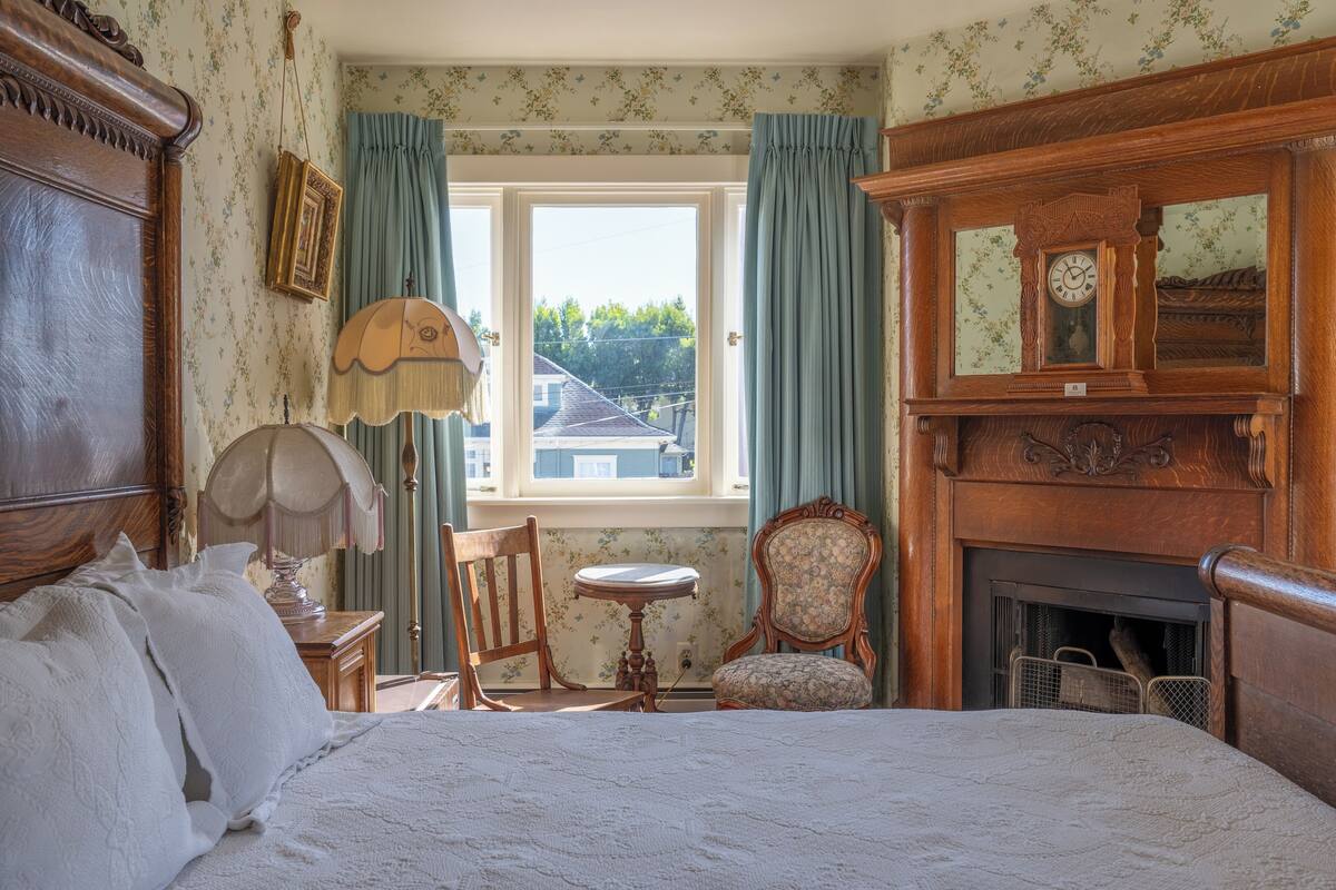 Ein Schlafzimmer mit einem Bett, einem Holzstuhl, einem kleinen Tisch, einer Lampe, einem Kamin und einem Fenster mit Vorhängen.