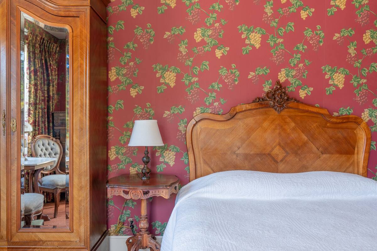 Ein Zimmer mit einem Bett, einem Nachttisch aus Holz mit einer Lampe, einem Sessel und einem Spiegel, der einen Essbereich widerspiegelt.