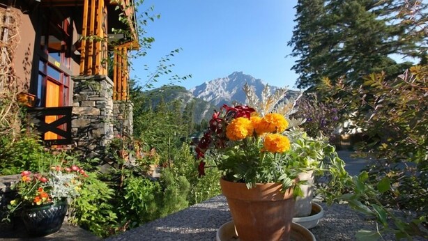 A rustic building with stone walls and wooden beams, surrounded by lush gardens and a mountain backdrop. - Buffaloberry B&B (Banff)
