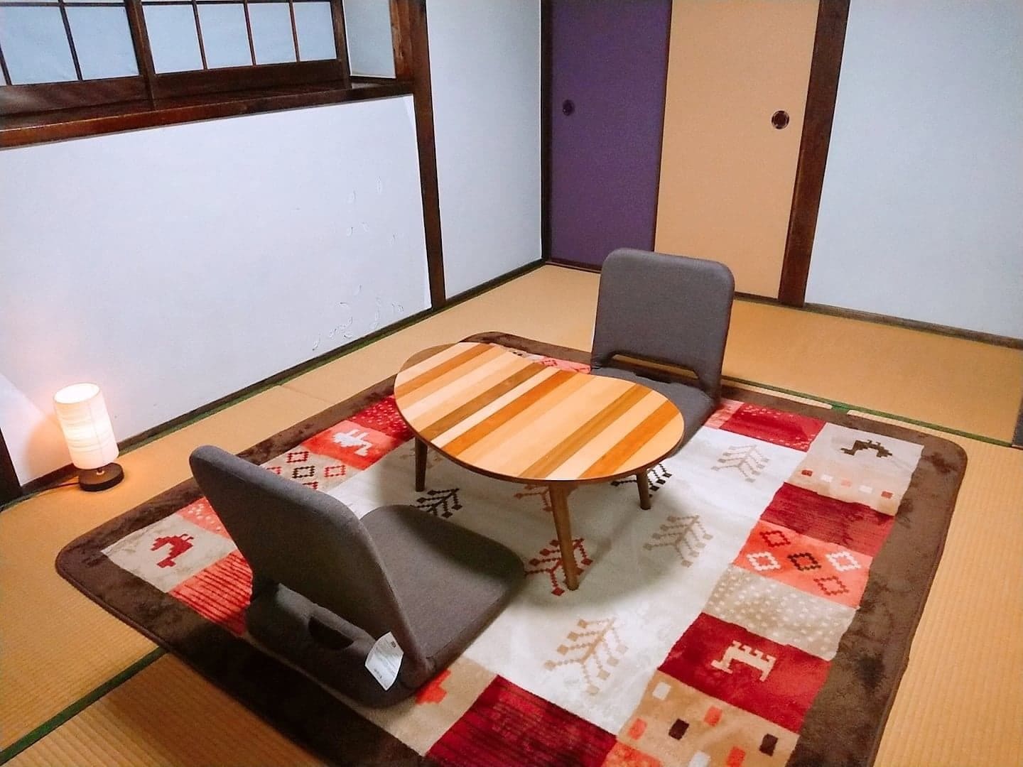 Una habitación con esteras de tatami, una mesa de madera baja, dos sillas y una lámpara de mesa pequeña.