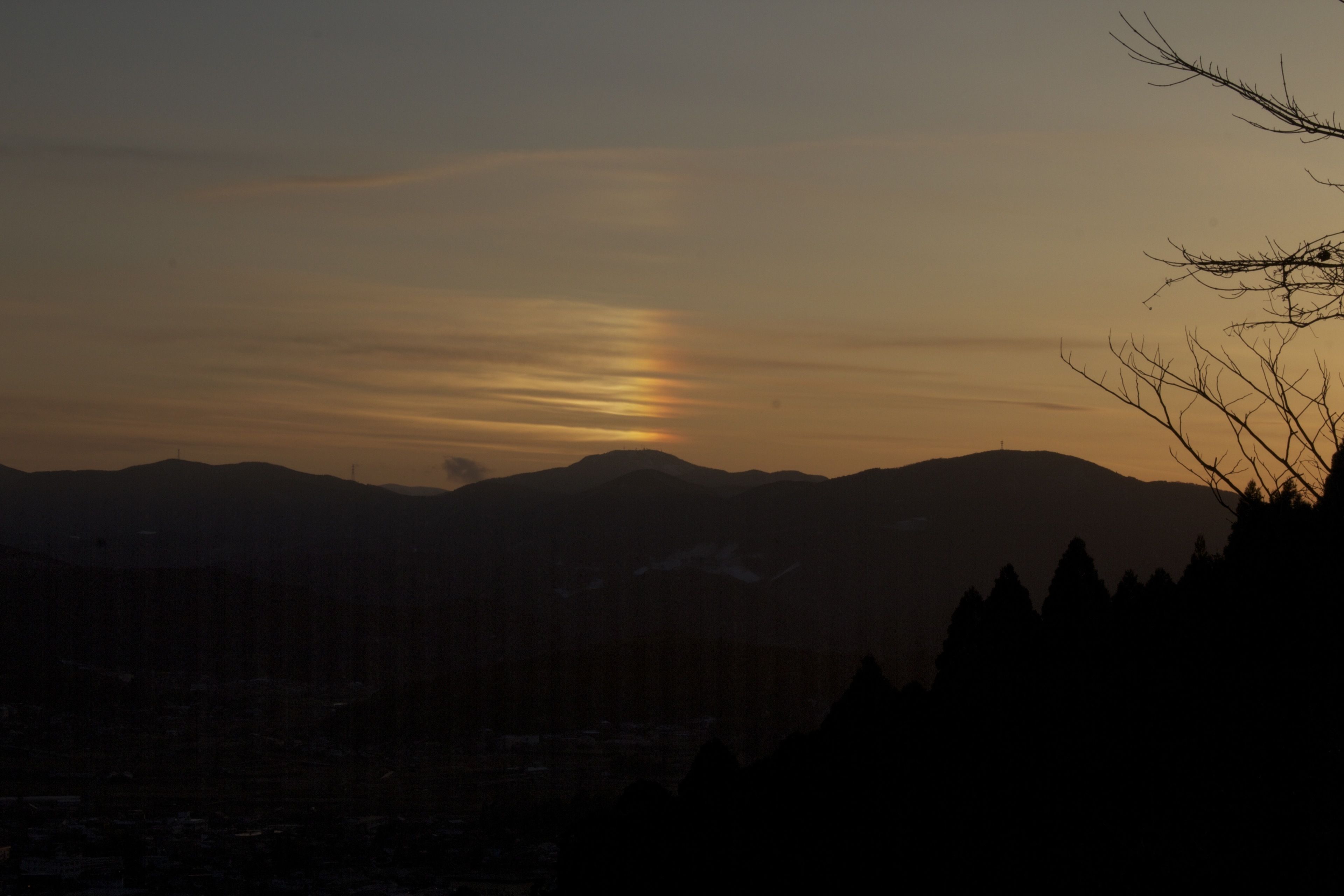 Un paysage de montagne au coucher du soleil, avec les silhouettes d’arbres et une ville en contrebas.