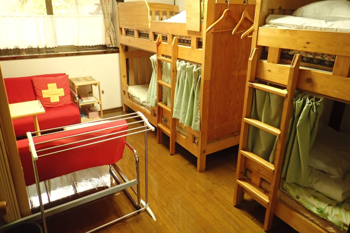 Une chambre de dortoir avec des lits superposés, un canapé rouge orné d’un coussin en forme de croix, un étendoir et une petite table en bois.