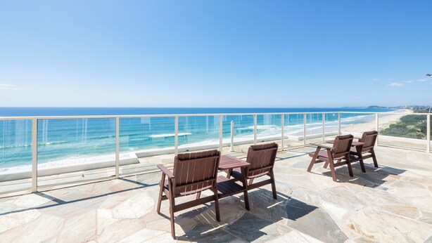 A terrace with stone flooring and wooden chairs overlooking a beach with waves and a clear sky. - Viscount on the Beach (Surfers Paradise)