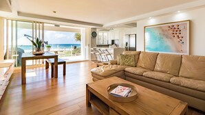 A spacious living room with a large leather sofa, wooden coffee table, and a dining area with a table and chairs. A kitchen is visible in the background, and there is a large window offering a view of the ocean. - Viscount on the Beach (Surfers Paradise)