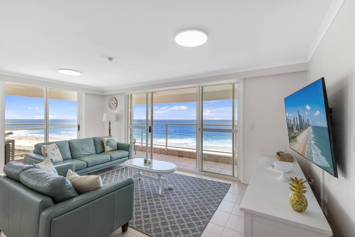A modern living room with a large window overlooking the beach, a sofa, a coffee table, and a flat-screen TV.