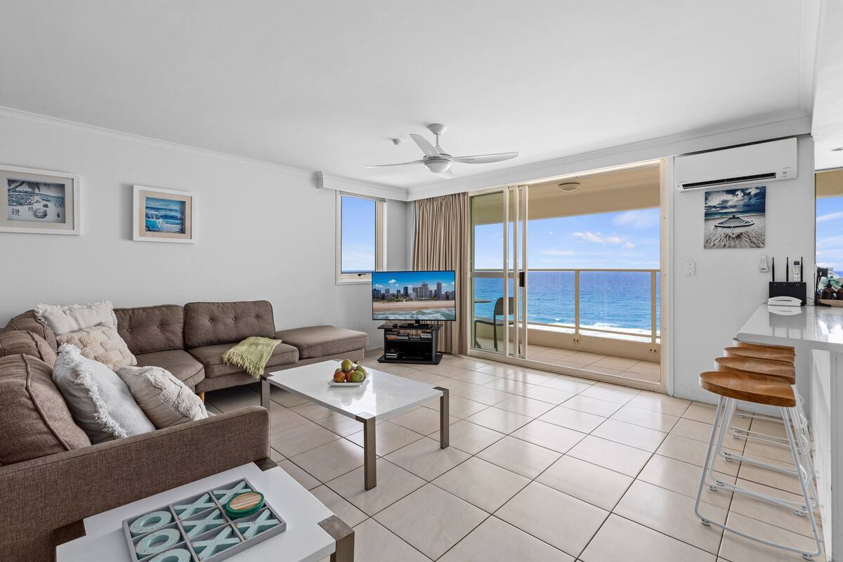 A modern living room with a sofa, coffee table, and TV, overlooking a balcony with a view of the sea.