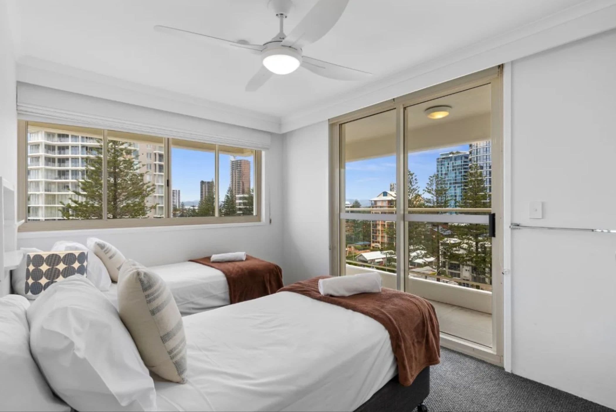 A hotel room with two beds, a ceiling fan, large windows with city views, and a sliding glass door.