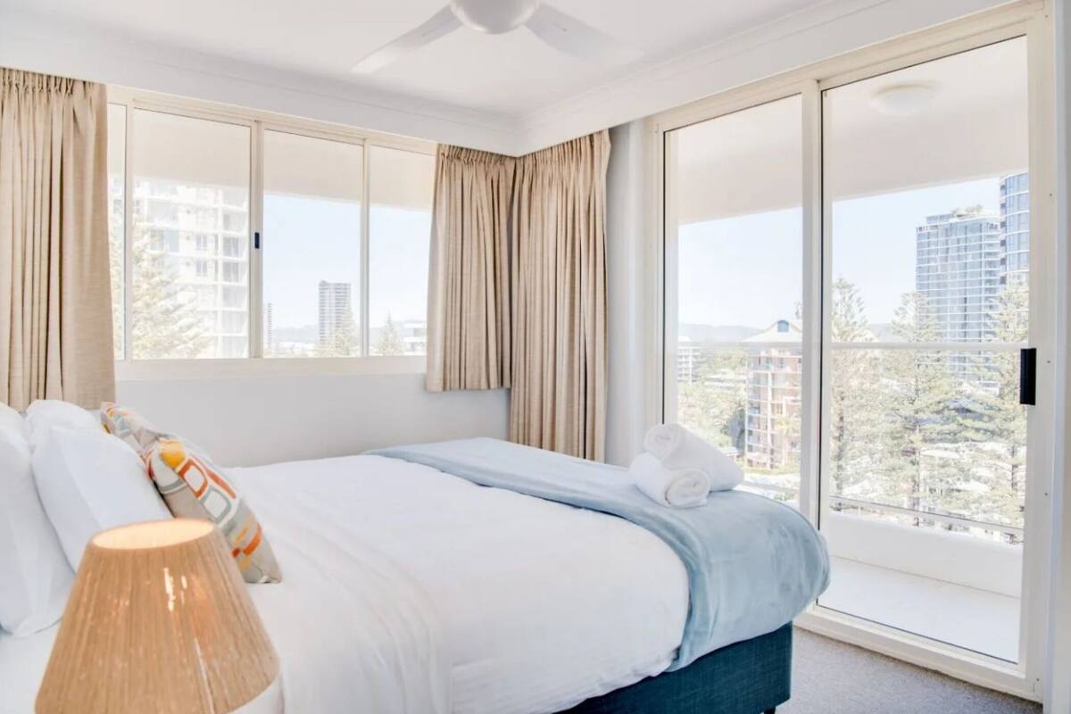 A hotel room with a large bed, a ceiling fan, and a view of the city through floor-to-ceiling windows.