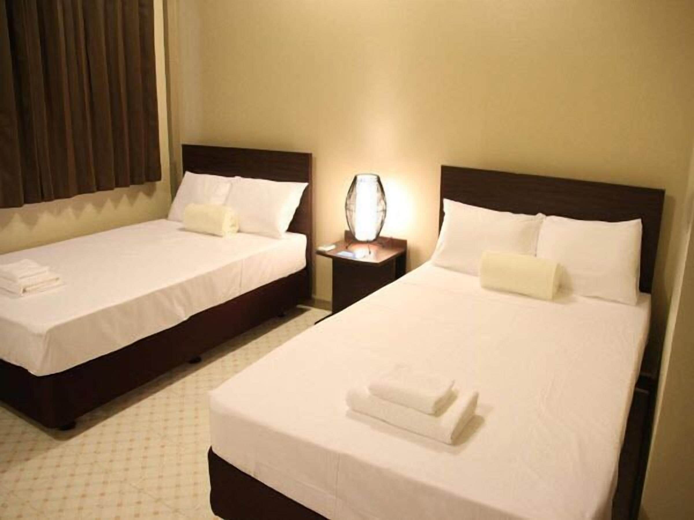 Dos camas individuales con sábanas blancas, una mesita de noche con lámpara entre ellas, y una ventana con cortinas.