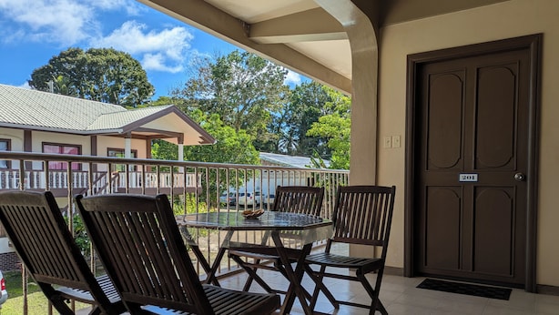 A balcony with four wooden chairs and a glass table, a door with the number 201, and a view of a building and trees in the background. - DW Motel Palau (Koror)