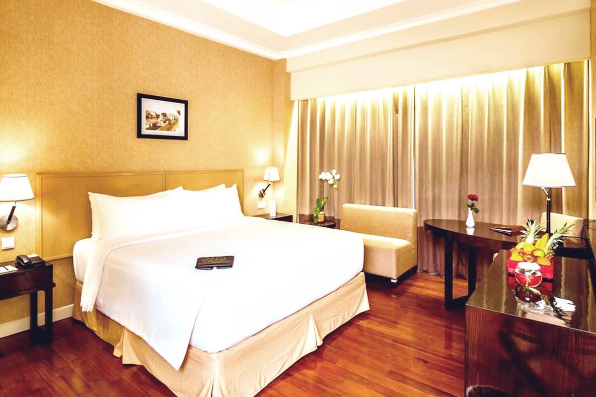 A hotel room with a large bed, a bedside table with a lamp, a chair, a small table with a fruit bowl, and a mirror.