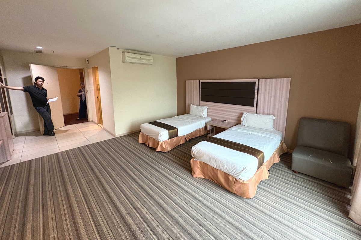 Ein Hotelzimmer mit zwei Betten, einem Sessel, einem Schreibtisch und einer Tür zu einem weiteren Raum.