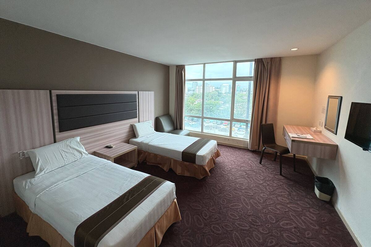 Ein Hotelzimmer mit zwei Betten, einem Schreibtisch, einem Stuhl und einem großen Fenster mit Blick auf die Stadt.