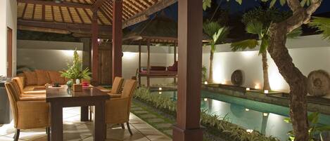 A covered outdoor seating area with a pool, wooden furniture, and a decorative tree.
