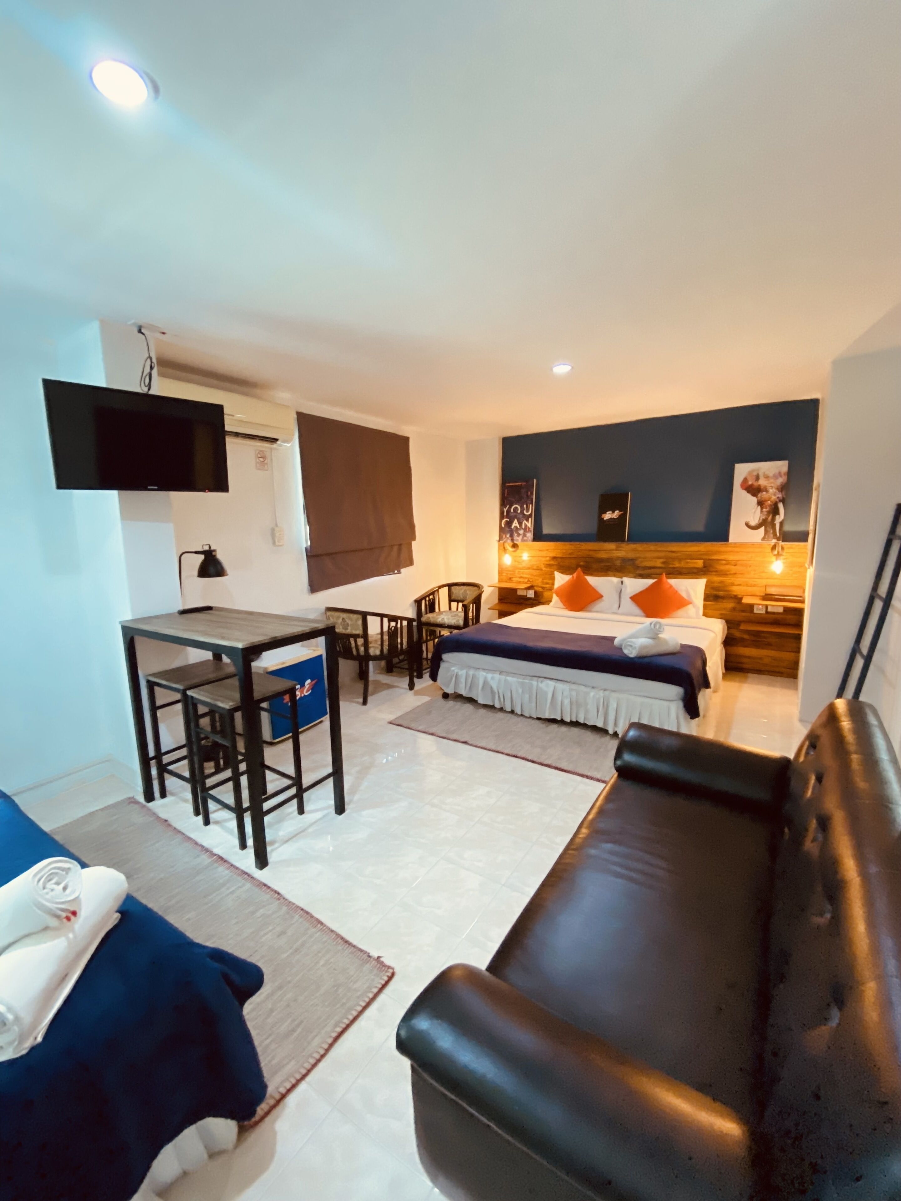 A compact living space with a sofa, a small dining table with chairs, a bed with pillows, a TV mounted on the wall, and a wooden accent wall.
