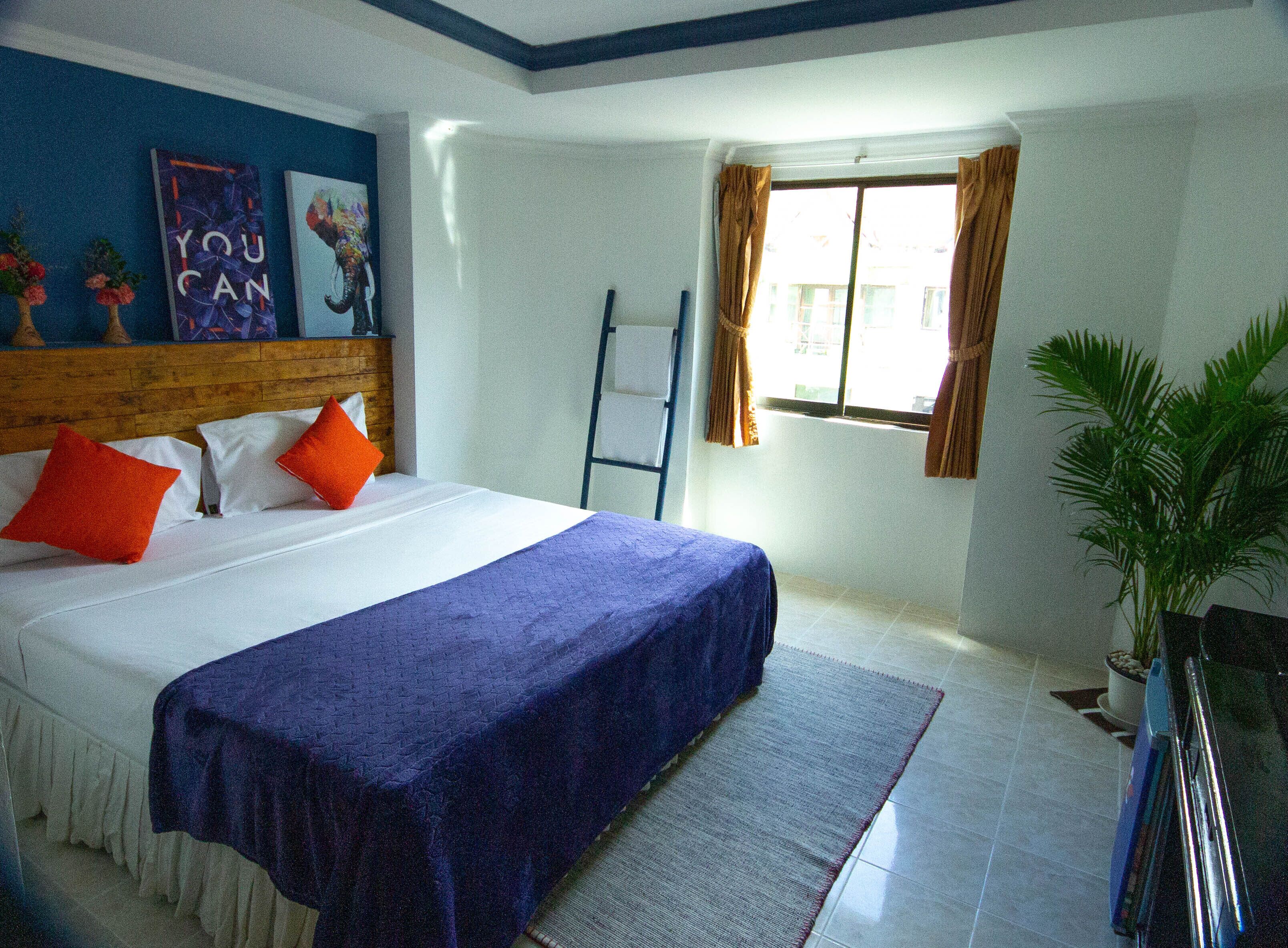 A bedroom with a bed, pillows, a blue bedspread, a window with curtains, a plant, and a ladder.