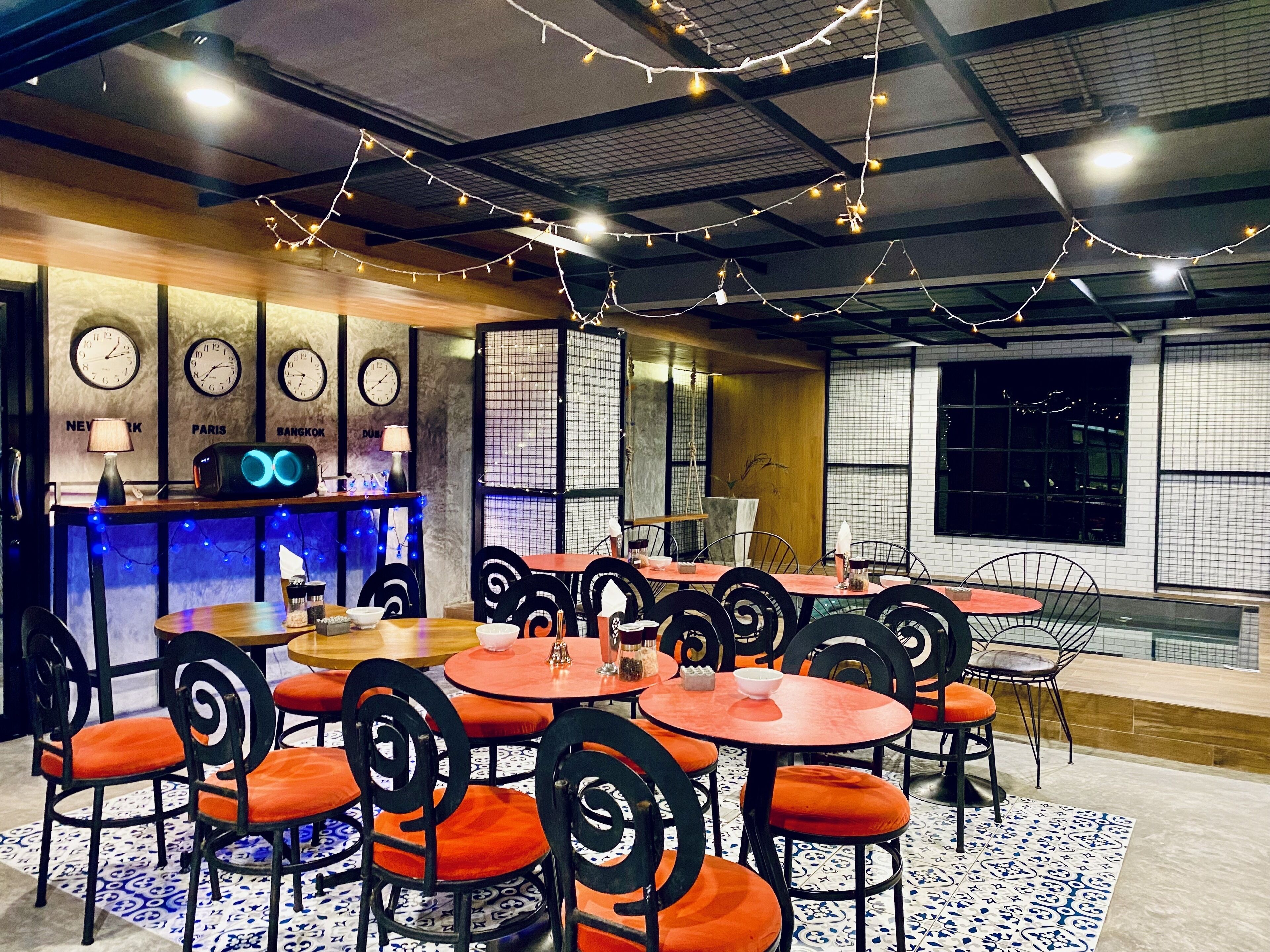 A restaurant with round tables and black chairs, a bar area with clocks, and string lights on the ceiling.